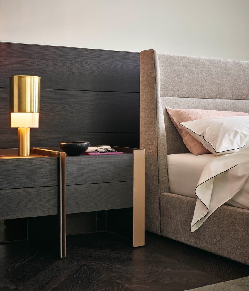 Chloe bed, padded headboard and bed frame with removable covering in fabric Naxos 07 cemento, piping in colour acciaio. Coordinated bed linen in cotton Provence 01 bianco with profile in nero. Blanket Linus granata. Quid wall panelling in black elm. Chloe night tables with outer structure in hide 29 tortora and inner structure in glossy brown nickel, drawers and top in black elm.