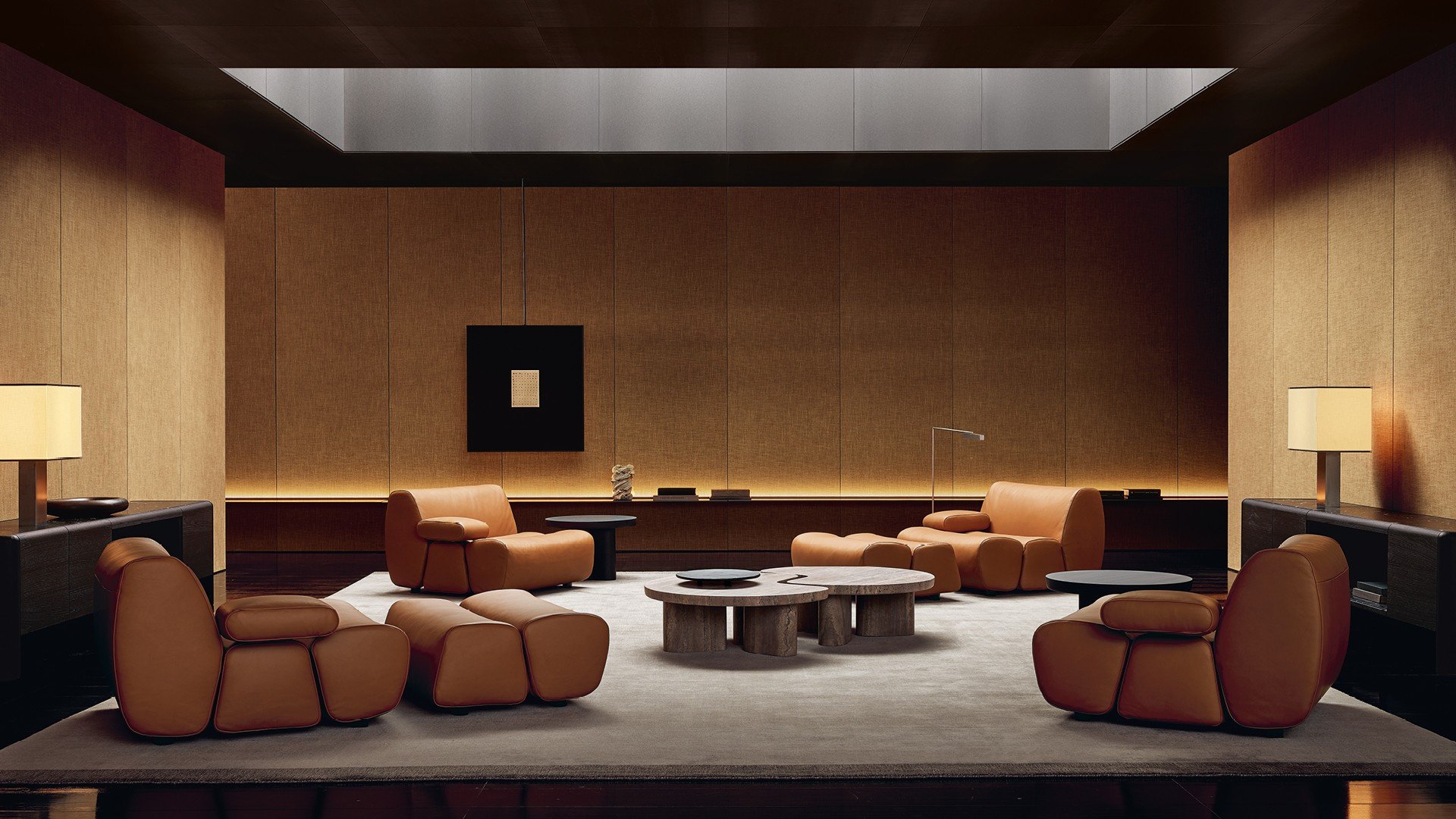 Ernest armchairs and chaise longue in removable leather Natural 05 cognac. Ernest coffee tables in mat marble travertino titanium and in black elm. Surface wall panelling in fabric Mesh bronzo, profiles in mat ardesia. Code backlit shelves in metal lacquer bronzo. Duke sideboard in black elm with top and open compartment lower shelf in mat marble nero marquina with polyester finish. Arthur floor lamp in satin-finish stainless steel and dimmable led light. Helga table lamps in satin-finish stainless steel and lampshade in white linen, dimmable led light. Carpet Plain tortora.