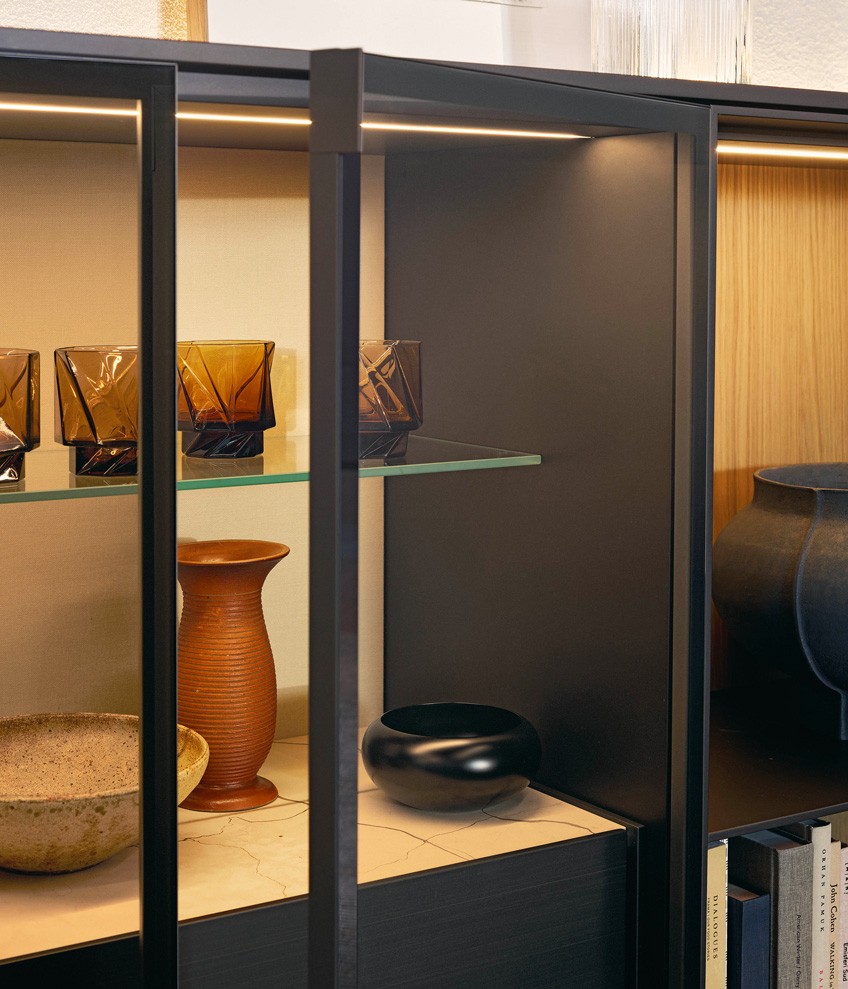 Detail of Code wall-mounted unit system with doors in transparent reflective glass, frame and handles with matt slate finish. Walls finished in light linen textile. Internal structure with bronze matt lacquer finish. Integrated LED lighting. Internal transparent glass shelves and drawers with bronze metal lacquer finish. Top in polished Zecevo marble. Backlit top with bronze metal lacquer finish.