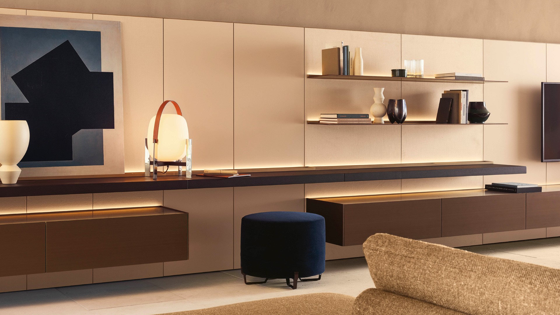Code composition with decorative panels covered in light linen textile. Profiles with matt champagne finish. Wall-mounted storage units with brass metal lacquer finish. Backlit top in polished Zecevo marble. Backlit shelf in black elm with cable channel in black elm and matt champagne profiles. Backlit shelves with brass metal lacquer finish. Westside sofa with removable cover in bronze Arcadia fabric (05). Base with Brown Nickel matt nickel finish. Westside coffee tables. Top in polished Sahara Noir marble with polyester finish and black elm. Structure in black elm. Kay Lounge armchair and ottoman with non-removable cover in roccia Silk leather (06). Chrome structure with satin finish. New York ottoman with removable cover in notte Tebe velvet (08), legs with Brown Nickel glossy nickel finish. Plain carpet in tortora.