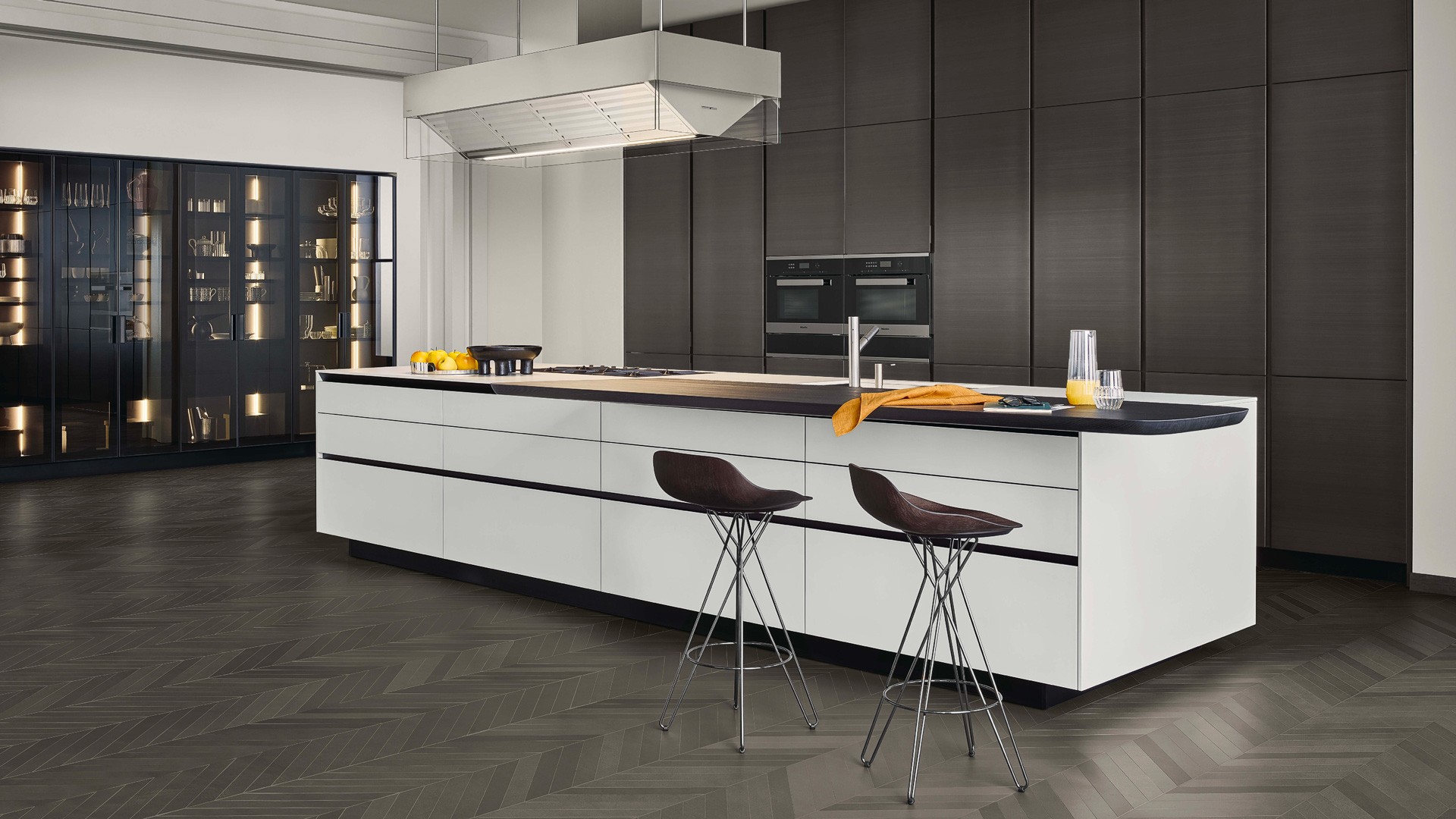 Base units with bianco extralight frosted glass doors, drawers and pull-out storage baskets with embossed lacquered carbone profile, sides and top in dekton bianco. Work island-hood in scotch brite steel and glass. Tall units with doors in metal lacquer bronzo and tall units PR22 with fumé glass doors. On the base units is the Sydney snack top in solid wood stained black elm. Harmony stools with seats in spessart oak and black chrome structure.
