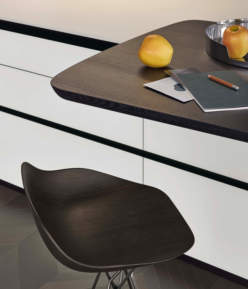 Base units with doors, drawers and pull-out storage baskets in bianco extralight frosted glass, sides and top in dekton bianco. On the base units is the Sydney snack top in solid wood stained black elm. Harmony stool with seat in spessart oak and black chrome structure.