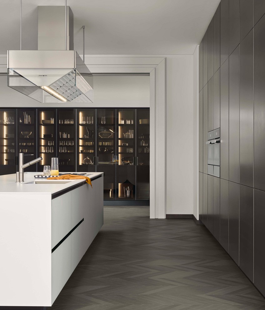 Base units with doors, drawers and pull-out storage baskets in bianco extralight frosted glass with embossed lacquered carbone profile, sides and top in dekton bianco. Work island-hood in scotchbrite steel and glass. Tall units with doors in metal lacquer bronzo and tall units PR22 with fumé glass doors, black anodized profile and integrated handle painted black.