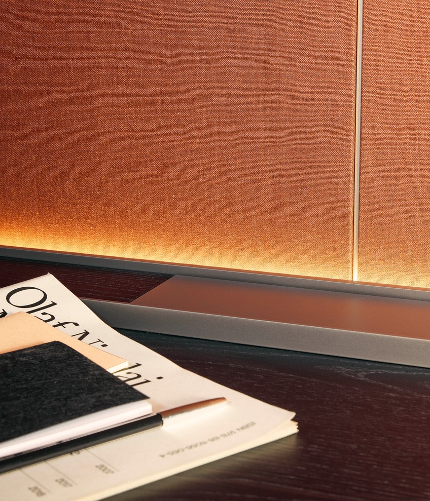 Деталь полки с задней подсветкой из черного вяза, канал с кабельными вводами из черного вяза с профилями с матовой отделкой цвета ардезия. Декоративные панели с покрытием из льняного текстиля цвета бручато. Профили с матовой отделкой цвета шампанское.