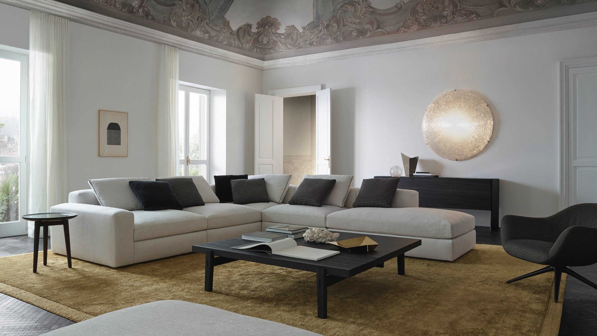 Sofa and pouf Dune with removable cover in fabric Rabat 400 ecru, cushions in fabric Nancy 101 carbone, velvet Persia 1404 carbone and fabric Rabat 400 ecru with border in grosgrain nero. Mad Chair armchair with removable cover in fabric Nancy 101 carbone, mat brown nickel swivel base. Carpet Frame gold.